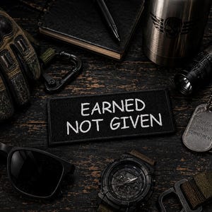 May include: A rectangular black embroidered patch with white text that reads EARNED NOT GIVEN, surrounded by tactical gear including a black glove, sunglasses, a wristwatch, a metal tumbler, a flashlight, and two metal dog tags with engraved text.