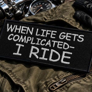 Puede incluir: Parche rectangular negro con texto bordado en blanco: "WHEN LIFE GETS COMPLICATED - I RIDE". El parche está sobre una chaqueta verde, con piezas de motocicleta, gafas de sol y un guante de cuero negro en el fondo.