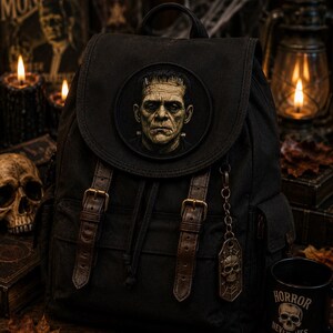 May include: A black canvas backpack featuring a circular patch of Frankenstein's monster on the front flap. The bag includes two brown faux leather straps with brass-toned buckles, a drawstring closure, and a hanging metal skull charm on a chain.