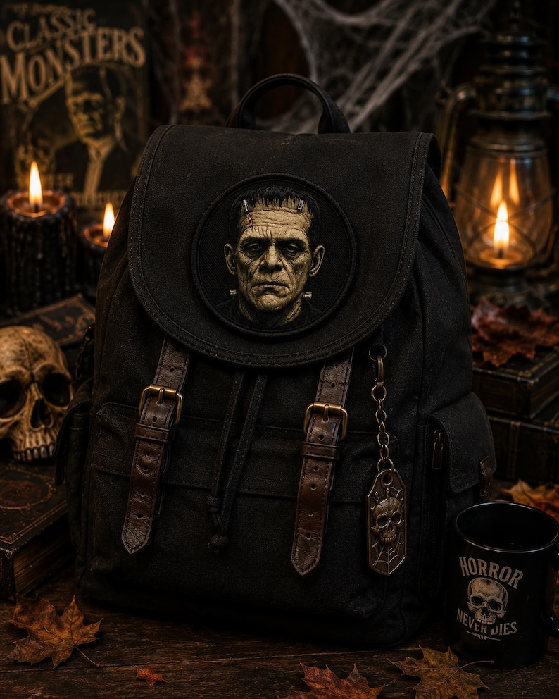 May include: A black canvas backpack featuring a circular patch of Frankenstein's monster on the front flap. The bag includes two brown faux leather straps with brass-toned buckles, a drawstring closure, and a hanging metal skull charm on a chain.