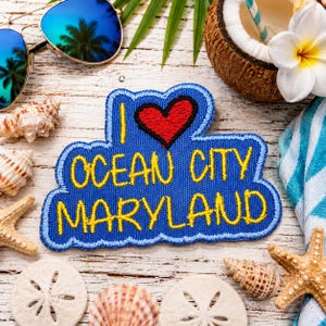 May include: A blue embroidered patch with a red heart and yellow text that reads I LOVE OCEAN CITY MARYLAND. The patch is surrounded by seashells, a starfish, blue-tinted sunglasses, a coconut shell, and a white tropical flower.