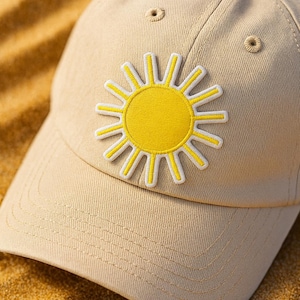 May include: A beige baseball cap with a yellow sun patch. The sun design has a yellow center and white-outlined yellow rays. The cap has a curved brim and a button on top.