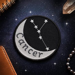 以下が含まれることがあります： 黒い背景に銀色の三日月と黒い刺繍でCancerという文字が施された円形の刺繍ワッペン。5つの星からなる銀色の星座模様が、月と黒い空間の上に配置されています。