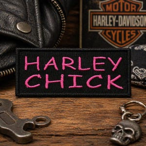 Puede incluir: Un parche rectangular negro con las palabras "HARLEY CHICK" bordadas en rosa. El parche se muestra sobre una superficie de madera, con una chaqueta de cuero y el logotipo de Harley-Davidson en el fondo. También se ve un llavero de calavera.