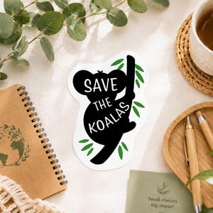 May include: A white sticker with a black koala silhouette holding onto a branch, with the words "SAVE THE KOALAS" in white. The sticker is surrounded by green leaves. A brown spiral notebook and a cup of tea are also visible.