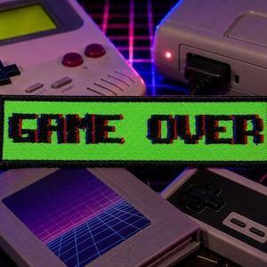 May include: A neon green patch with the words "GAME OVER" in pixelated red and blue lettering. The patch is surrounded by retro gaming consoles, including a handheld device, a cartridge, and a controller, evoking a nostalgic gaming theme.