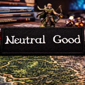 May include: A black patch embroidered with the words "Neutral Good" in white. The patch is set against a backdrop of dice, miniature figures, and books, suggesting a tabletop gaming theme. The dice are various colors and shapes.