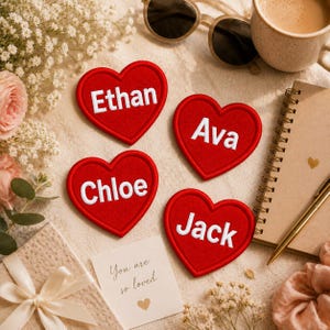May include: Four red heart-shaped patches with white embroidered names: Ethan, Ava, Chloe, and Jack. A small card reads "You are so loved." The image includes a notebook, pen, sunglasses, and a cup of coffee, with floral accents.