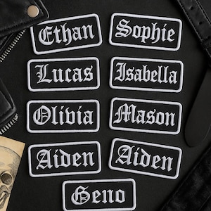 May include: A collection of black rectangular patches with white embroidered names in a gothic font. Names include Ethan, Sophie, Lucas, Isabella, Olivia, Mason, Aiden, and Geno. The patches are displayed on a black leather surface.