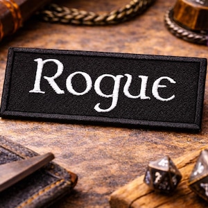 Może przedstawiać: Czarna naszywka z białym napisem "Rogue". Obraz zawiera kości do gry i skórzane akcesoria, sugerując motyw gry fabularnej, na drewnianej powierzchni.