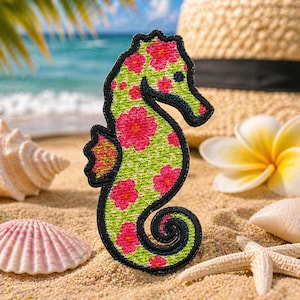May include: Embroidered seahorse patch with a green background and pink floral pattern, outlined in black. The patch is on a sandy beach with a straw hat, seashells, and a starfish in the background. The seahorse is a tropical theme.