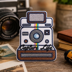 May include: Embroidered patch in the shape of a vintage instant camera. The patch is white with a gray lens and flash, and a brown border. The text "Capture Life" is written in cursive. A colorful stripe runs down the center. Photography theme.