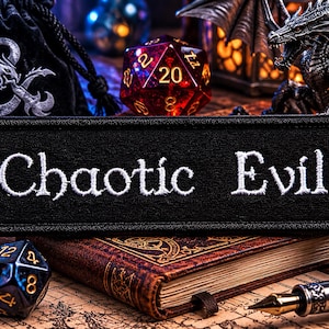 Könnte beinhalten: Ein schwarzer Aufnäher mit den weißen Worten "Chaotic Evil", der auf einem Buch liegt. Das Bild zeigt verschiedene Würfel, eine Drachenfigur, eine D&D-Tasche und eine brennende Kerze, was auf ein Tabletop-Gaming-Thema hindeutet.