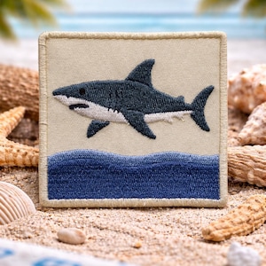 May include: Embroidered patch featuring a dark blue shark swimming above blue waves. The shark is set against a beige background, with a beige border. The patch is square-shaped and suitable for application on clothing or accessories.