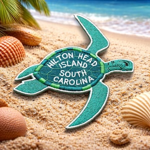 May include: A teal embroidered patch shaped like a sea turtle, with the text "Hilton Head Island South Carolina" in white. The patch is on a sandy beach with seashells and ocean in the background.