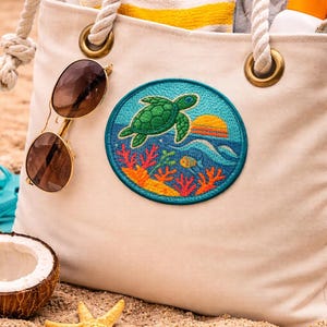 May include: A cream-colored beach tote bag with a turtle patch, sunglasses, and a yellow and white striped towel. The patch features a green turtle, coral, and a sunset. A coconut, starfish, and blue flip-flops are on the sand.