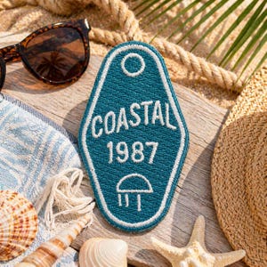 May include: A teal patch with the word "COASTAL" and the year "1987" embroidered in white. The patch is shaped like a hotel key tag and is surrounded by beach-themed items, including sunglasses, a straw hat, and seashells.