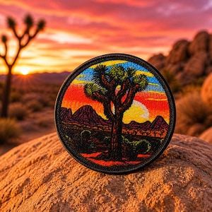 May include: A circular patch featuring a Joshua tree silhouette against a sunset backdrop. The patch has a black border and showcases vibrant colors of orange, red, yellow, and blue. The design includes mountains and a setting sun, evoking a desert landscape.
