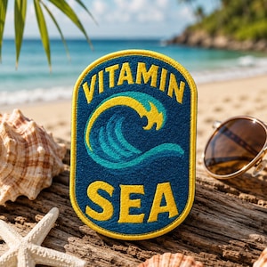 May include: A navy blue patch with the words "VITAMIN SEA" in yellow, and a yellow and teal wave design. The patch is on driftwood, with seashells and sunglasses in the background. The beach and ocean are visible.