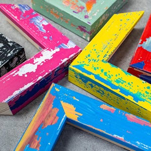 Può includere: Assortimento di angoli di cornici in legno colorati e usurati. Le cornici sono dipinte in vari colori tra cui rosa, giallo, rosso, blu, nero e verde, con schizzi di vernice bianca. Le cornici sono disposte in un motivo circolare.