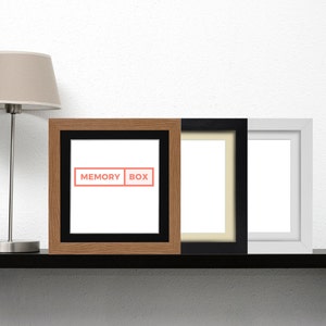 Könnte beinhalten: Drei Bilderrahmen auf einem schwarzen Regal. Der Rahmen links ist aus Holz mit einer schwarzen Passepartout und dem Text "MEMORY BOX". Der mittlere Rahmen ist schwarz mit einer cremefarbenen Passepartout. Der Rahmen rechts ist weiß.