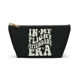 Könnte beinhalten: Schwarze Kosmetiktasche mit Reißverschluss und dem Text "IN MY FLIGHT ATTENDANT ERA" in Weiß mit einer Retro-Schriftart und Flugzeugsymbolen.