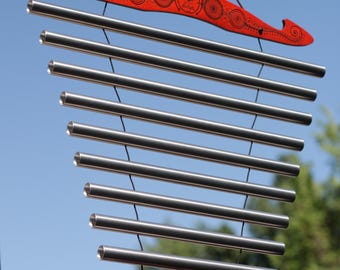 May include: A wind chime with a red wooden top and bottom piece, each with a decorative design. The chime has multiple silver metal tubes suspended from the top piece. The background is a clear blue sky.