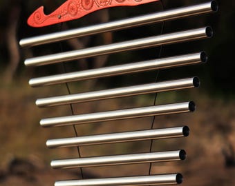 May include: A wind chime with a red wooden top and bottom, featuring etched circular designs and the word "divya". The chime has multiple silver metal tubes suspended by black strings, creating a soothing sound.