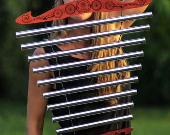 May include: A woman holds a wind chime with silver tubes and red wooden accents. The wind chime has a decorative red top and bottom piece. The background is a blurred green outdoor scene.