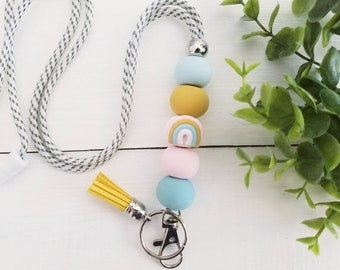 May include: A white and gray lanyard with a yellow tassel and a key ring. The lanyard has a string of colorful beads, including blue, yellow, pink, and a rainbow bead.