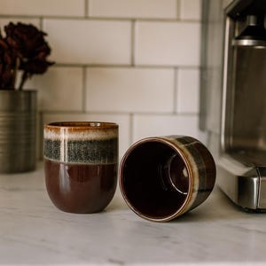 May include: Two ceramic coffee mugs with a dark brown base and a band of brown, gray, and cream near the rim. One mug is upright, the other is on its side. The mugs are on a white countertop.