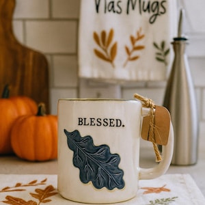 Puede incluir: Taza de cerámica color crema con la palabra "BLESSED" y un diseño de hoja azul. Una etiqueta marrón cuelga del asa. Una toalla a juego con las palabras "Nas Mugs" y diseños de hojas de otoño está en el fondo. También se ven dos calabazas naranjas.