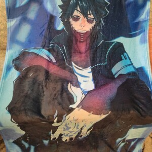 May include: A plush blanket featuring a detailed anime character illustration. The character has dark hair, stitched facial features, and is wearing a black jacket over a white shirt. The background has blue and purple hues.