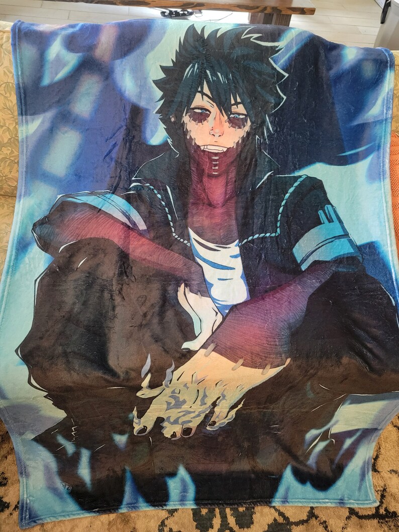 May include: A plush blanket featuring a detailed anime character illustration. The character has dark hair, stitched facial features, and is wearing a black jacket over a white shirt. The background has blue and purple hues.