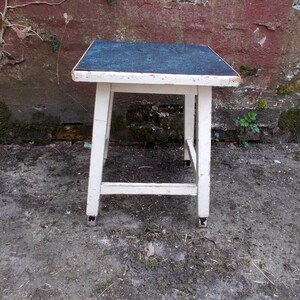 Könnte beinhalten: Ein weißer Holzhocker mit einer dunkelblauen Oberseite. Der Hocker hat ein einfaches Design mit vier Beinen und einer quadratischen Oberseite.