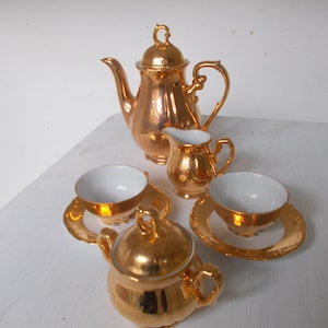 Könnte beinhalten: Ein Vintage-Teeservice mit metallisch goldener Oberfläche. Das Set enthält eine Teekanne, einen Sahnekännchen, eine Zuckerdose und zwei Teetassen mit Untertassen. Die Innenseiten der Tassen und des Sahnekännchens sind weiß.