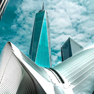 May include: A low angle view of a modern skyscraper with a sharp, pointed top, surrounded by other buildings with a white, geometric structure in the foreground.