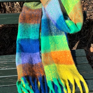 May include: A colorful scarf with a plaid pattern in shades of blue, green, orange, and yellow. The scarf has a soft, fuzzy texture and fringed ends. It is draped over a wooden bench.