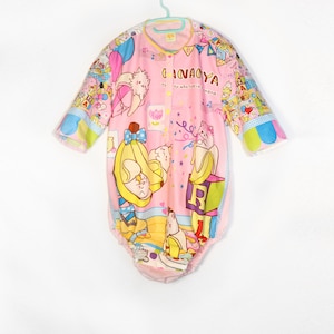 May include: Pink baby onesie with a colourful cartoon print of animals and musical notes. The onesie has a button closure and a ruffled hem. The text on the onesie reads "Chanaya The kitty who lives in a banana."