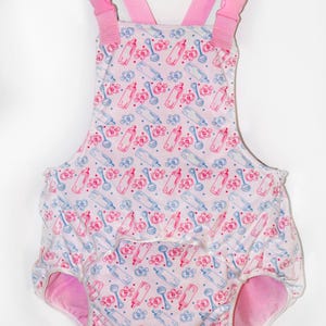 May include: A light pink romper featuring a repeating pattern of blue and pink baby bottles and pacifiers. The garment includes adjustable pink shoulder straps with plastic buckles and leg openings with pink fabric lining the interior edges.