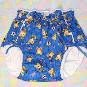 May include: Blue baby nappy with a tiger print. The nappy features a blue striped pattern with cartoon tigers, stars, and other graphics. White trim and ties are visible. The nappy is designed for babies and toddlers.