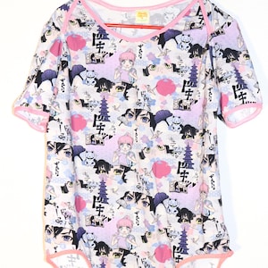 May include: A white onesie with a pink trim and short sleeves. The onesie is covered in a colourful anime-style pattern featuring characters, pagodas, and Japanese text. The design includes pink, blue, and black elements.