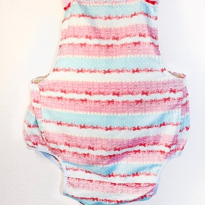 May include: A pink and blue striped baby overall with a white lace trim and pink straps. The overall has a pocket on the back and is made of a soft, comfortable fabric.