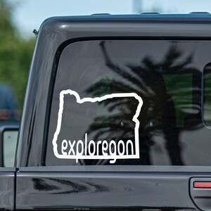 May include: White vinyl sticker on a black car window. The sticker is an outline of the state of Oregon with the text "exploreoregon" underneath.