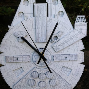 May include: A white wooden clock shaped like the Millennium Falcon spaceship. The clock has black hands and detailed engraved lines. The clock measures approximately 18 inches in diameter.
