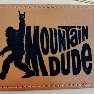 May include: A rectangular, brown leather patch with a black silhouette of Bigfoot raising a hand in a rock-and-roll gesture. The words "MOUNTAIN DUDE" are printed in black, stylized lettering.