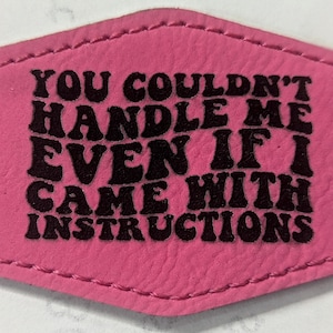 May include: A pink leather patch with black text that reads "YOU COULDN'T HANDLE ME EVEN IF I CAME WITH INSTRUCTIONS". The patch has a stitched border and a unique shape.