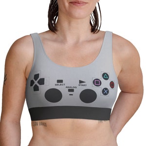 May include: A grey sports bra with a video game controller design. The bra has a black band at the bottom and a graphic of a controller with buttons and directional pad. The bra is sleeveless.