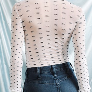 May include: A white sheer long-sleeved shirt with a black repeating pattern of the letter "F". The shirt is worn over a pair of dark blue jeans.
