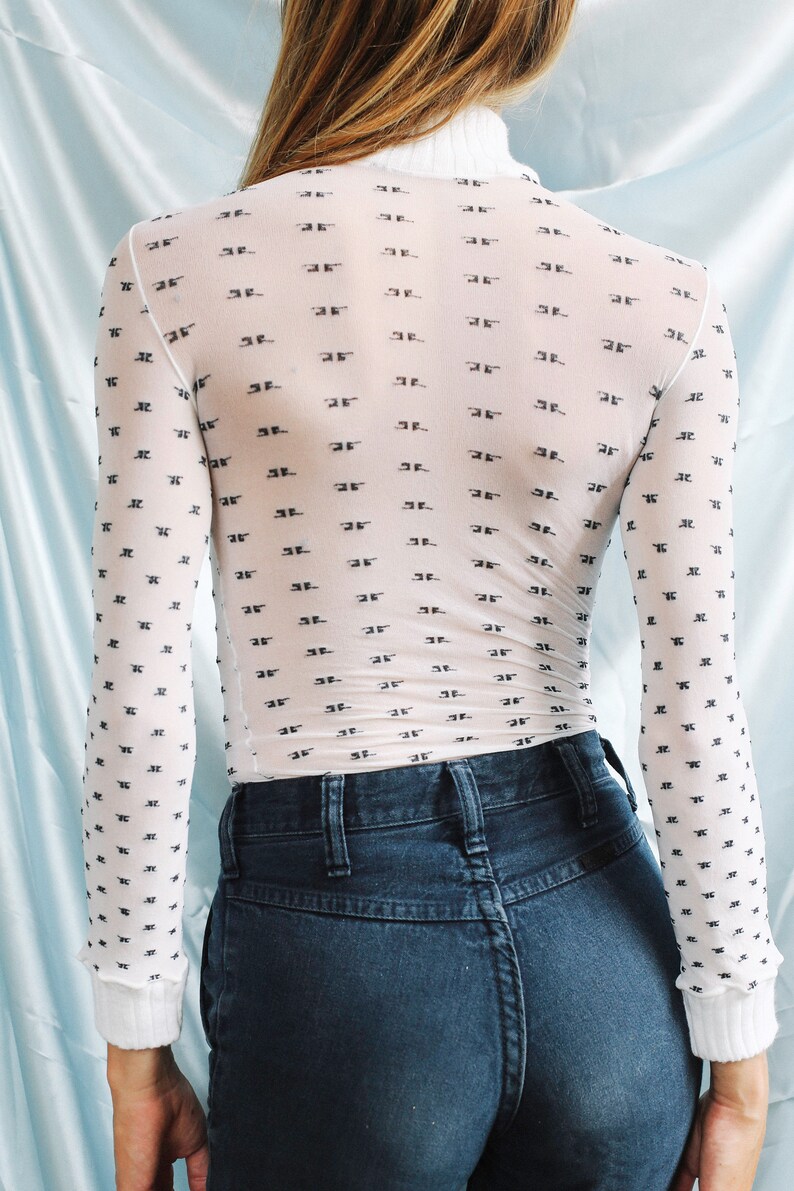 May include: A white sheer long-sleeved shirt with a black repeating pattern of the letter "F". The shirt is worn over a pair of dark blue jeans.